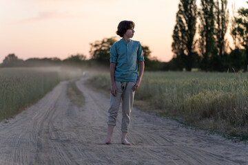 A barefoot teenager in comfortable clothes stands on a dirt road.