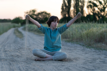 A barefoot teenager in comfortable clothes sits on a dusty dirt road.
