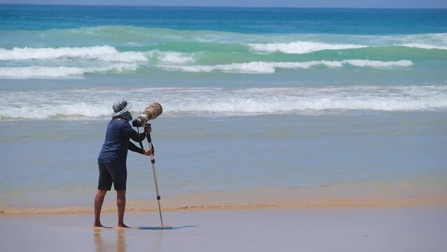 A photographer on a beautiful beach takes pictures of surfers with a camera with a large telephoto lens and a monopod.