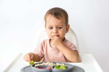 cute 1 year old baby sitting in a high chair and eating complementary foods on white background, baby feeding and childhood concept