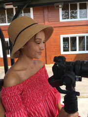 Asian girl in a wicker hat and red dress holds a camera on a stabilizer with a large lens - obrazy, fototapety, plakaty
