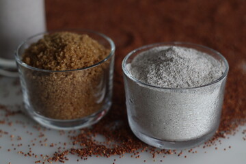 Ragi flour and raw sugar in a glass bowl along with a scattered spread of ragi grains around