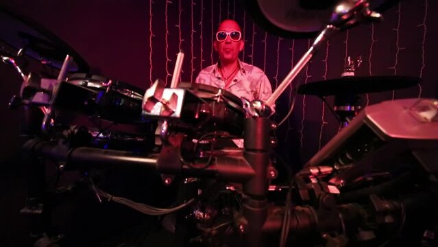 Bald man in sunglasses and white shirt plays drum set 
