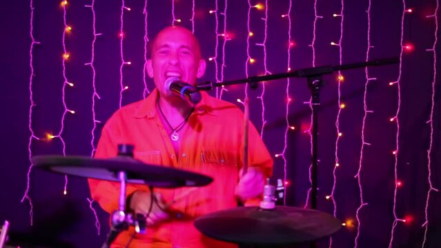 Bald man in orange shirt plays drum set and sings in night club