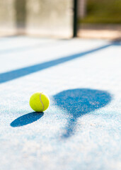 Vertical photo of a padel ball and the shadow of a racket on the floor of an outdoor court. Padel...