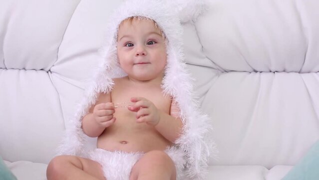 Cute little kid in white soft pants and hat sits on white sofa