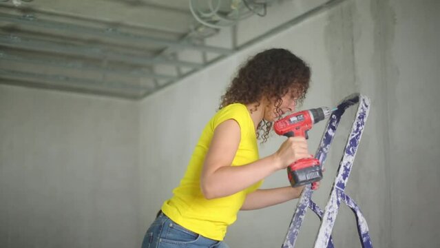 Woman with electric screwdriver ups on ladder in new apartment