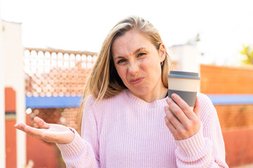 Young blonde woman holding a take away coffee at outdoors making doubts gesture while lifting the shoulders
