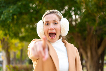 Young blonde woman wearing winter muffs at outdoors surprised and pointing front