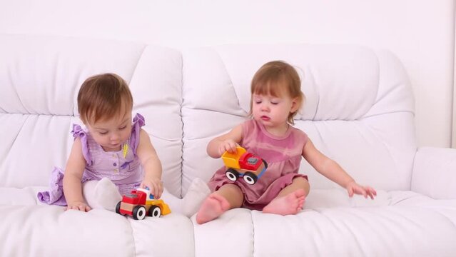 Two little girls in dresses sit on sofa and play with toy cars