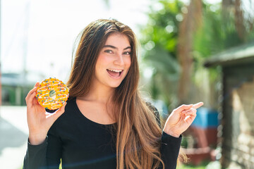 Young pretty caucasian woman holding a donut at outdoors surprised and pointing finger to the side