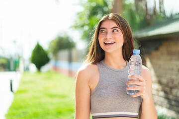 Young pretty caucasian woman with a bottle of water at outdoors looking up while smiling