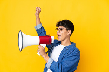 Young man over isolated yellow background shouting through a megaphone to announce something in...