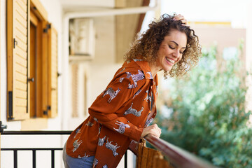 Smiling young woman looking down while standing on balcony of house - obrazy, fototapety, plakaty