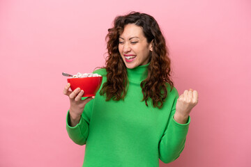 Young caucasian woman holding a bowl of cereals isolated on pink background celebrating a victory