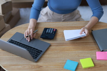 Asian woman starting a small business at home working with parcel boxes and calculating revenue and profit from sales.