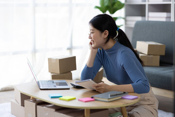 Asian woman starting a small business at home working with parcel boxes and calculating revenue and profit from sales.