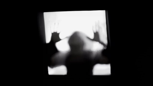 A scary shadow behind an opaque glass window, lurking while standing still, hands shaped like blades. Silhouette of a zombie or a serial killer. Horror vibes, flickering lights.
