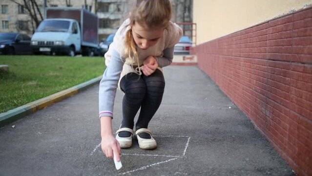 Little cute girl draws house on asphalt by white chalk on street