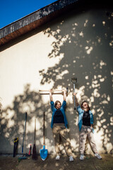 Two happy lesbian girls pose and laugh with garden tools near the wall in the shade of trees and the rays of the sun