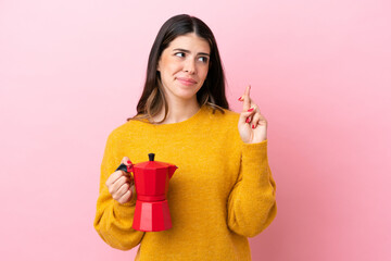 Young Italian woman holding a coffee maker isolated on pink background with fingers crossing and wishing the best