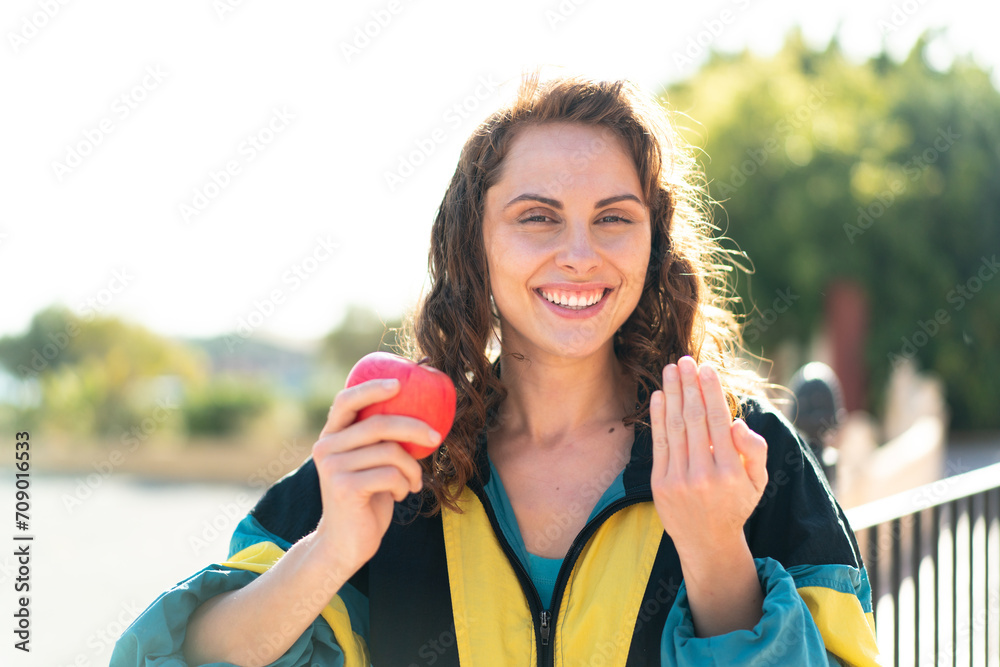 Wall mural Young sport woman with an apple at outdoors inviting to come with hand. Happy that you came