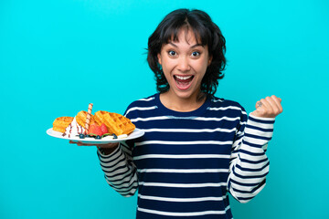 Young Argentinian woman holding waffles isolated on blue background celebrating a victory in winner position