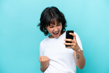 Young Argentinian woman isolated on blue background using mobile phone and doing victory gesture