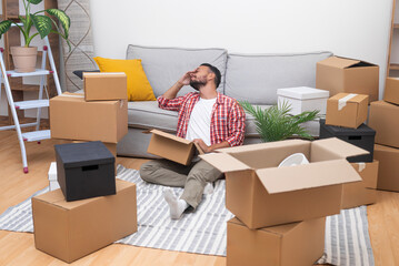 Smiling man in shirt with delighted expression sits on floor with carton box enjoying moving to...