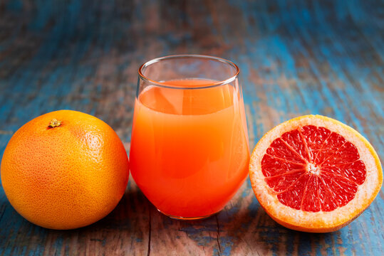 Glass of grapefruit juice next to a whole fruit and a half piece showing its reddish interior, rustic background.