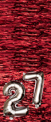 Silver foil balloon number number 27 on a background of red tinsel decoration. Birthday greeting...