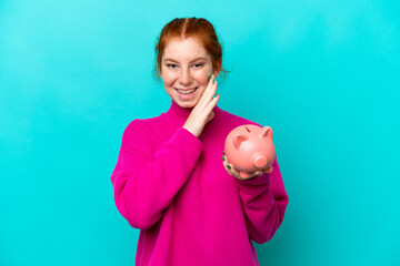 Young caucasian reddish woman holding a piggybank isolated on blue background whispering something