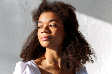 Tender portrait of young african american woman with curly hairstyle standing in sunlight indoors against grey wall - obrazy, fototapety, plakaty