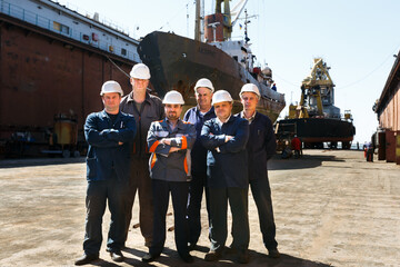 Maintenance crew poses in front of large vessel at shipyard dry dock. Engineers in hardhats,...