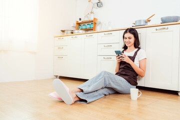 Happy young woman using smart phone relaxing at home She sitting on floor at kitchen using mobile phone Happy smiling Asian girl using smartphone for shopping online or chatting online with friend