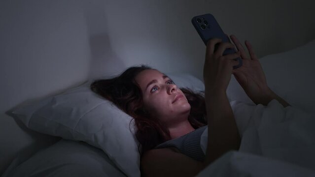A woman, in the dark of night, lies in bed, fixated on her phone's glow, image portraying the struggles of insomnia and smartphone addiction. 