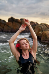Woman in black swimsuit enjoys serene swim in tropical lagoon. Red flower adorns her hair, symbol of exotic leisure. Calm water reflects scenic sunset, travelers peaceful retreat in nature.
