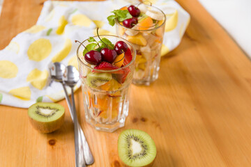 View from the top to large transparent glasses with chopped fruits and berries and long spoons on the table. Mandarin, kiwi, banana, strawberries, blueberries, cherries and mint leaves. Healthy snack.