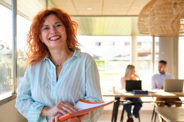 Redhead middle aged business woman looking at camera and smiling at coworking office.