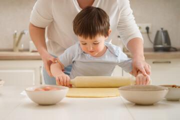 A mother woman teaches a toddler baby boy to roll out the dough with a pie rolling pin. Process of...