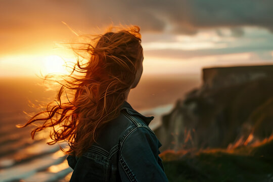 girl standing on a cliff, looking out at the sunset, with her hair blowing in the wind