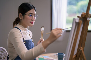 Young asian female artist hold paintbrush and use brush to drawing and painting artwork on canvas