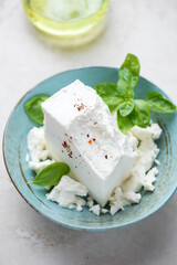 Turquoise bowl with a block of feta cheese and green basil, vertical shot on a light-beige stone background, selective focus
