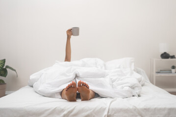 woman awakens in bed, bare feet and hand with cup of coffee out of blanket , stretching.