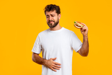 Upset Man Touching Aching Stomach Holding Burger Over Yellow Background