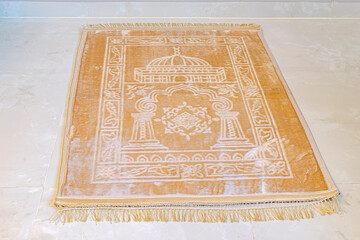 A muslim carpet set on the floor, beige brownish color with a mosque, curves and rectangle patterns. 
