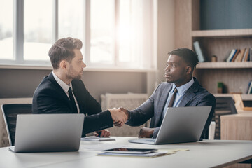 Two confident businessmen in formalwear shaking hands while sitting at the desk in modern office