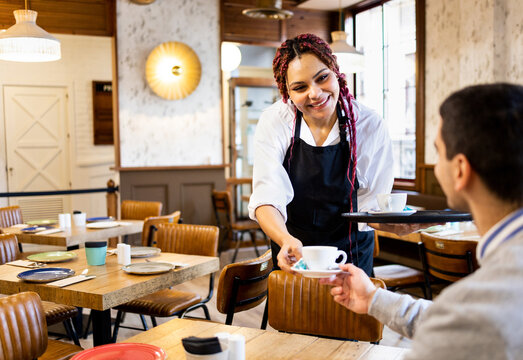 A dark-skinned woman works inside a cafeteria dressed in a uniform. The African girl serves a cup of coffee to a client.Working woman concept.Women working in the hospitality industry.