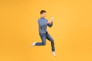 Fototapeta na wymiar Happy Excited Teen Boy With Smartphone In Hands Jumping On Yellow Background