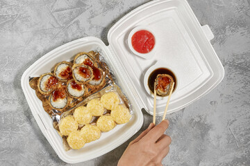 Delicious sushi rolls in disposable plastic boxes. The hand takes a roll with chopsticks. Food delivery service concept, flat stacking